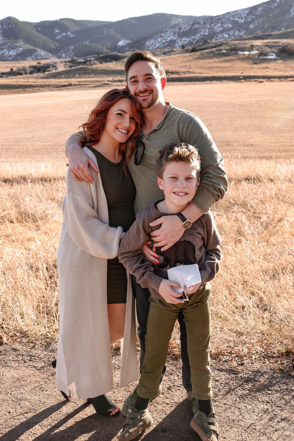 A family of three poses together in a golden field with mountains in the background.