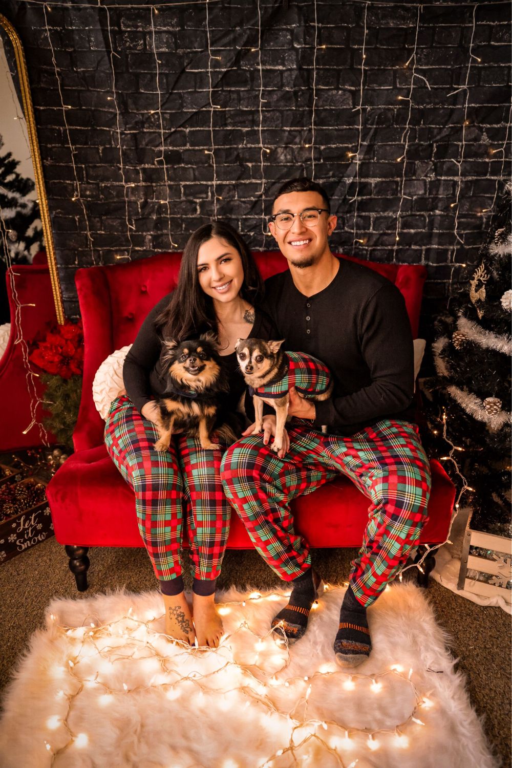 A couple in matching plaid pajamas sits on a red couch with two small dogs during Christmas.