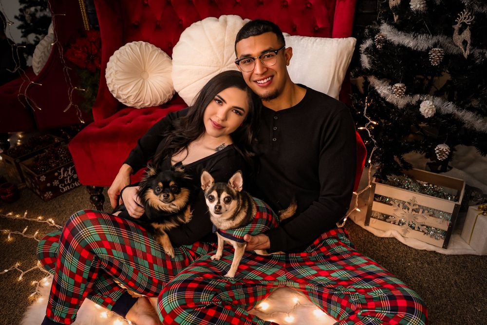 A cozy holiday portrait of a couple with their two dogs on a red sofa near a Christmas tree.