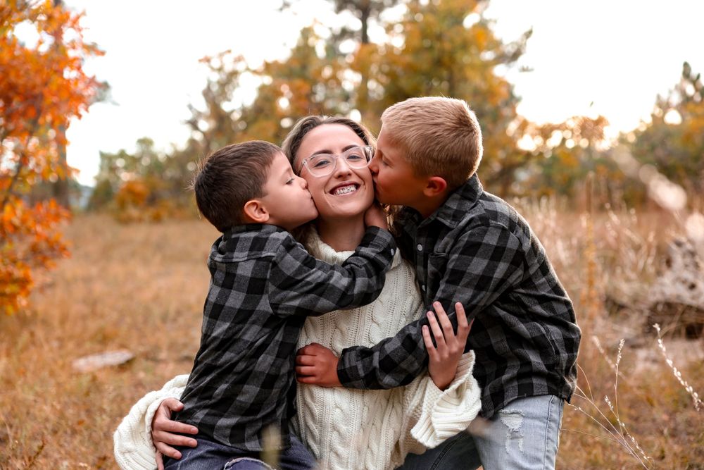 Children in plaid shirts give sweet kisses to someone wearing a white sweater in fall foliage.