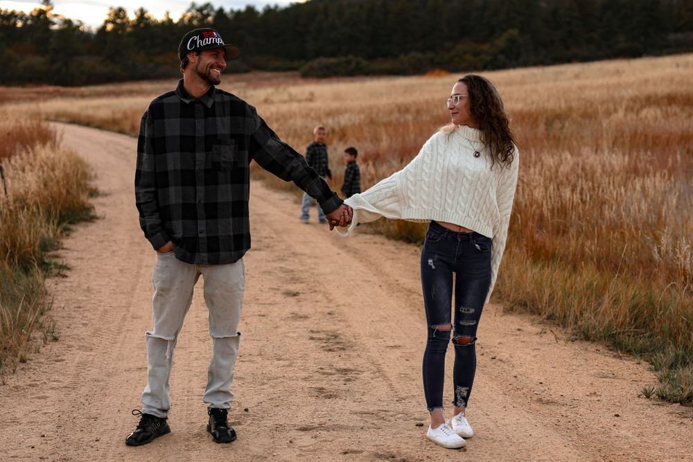A couple holds hands while walking on a dirt path through an autumn field at sunset.