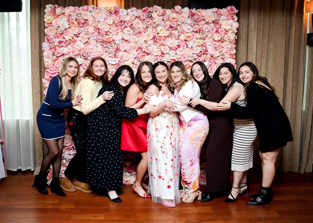 A group of women posing together in front of a pink floral backdrop at a celebratory event.