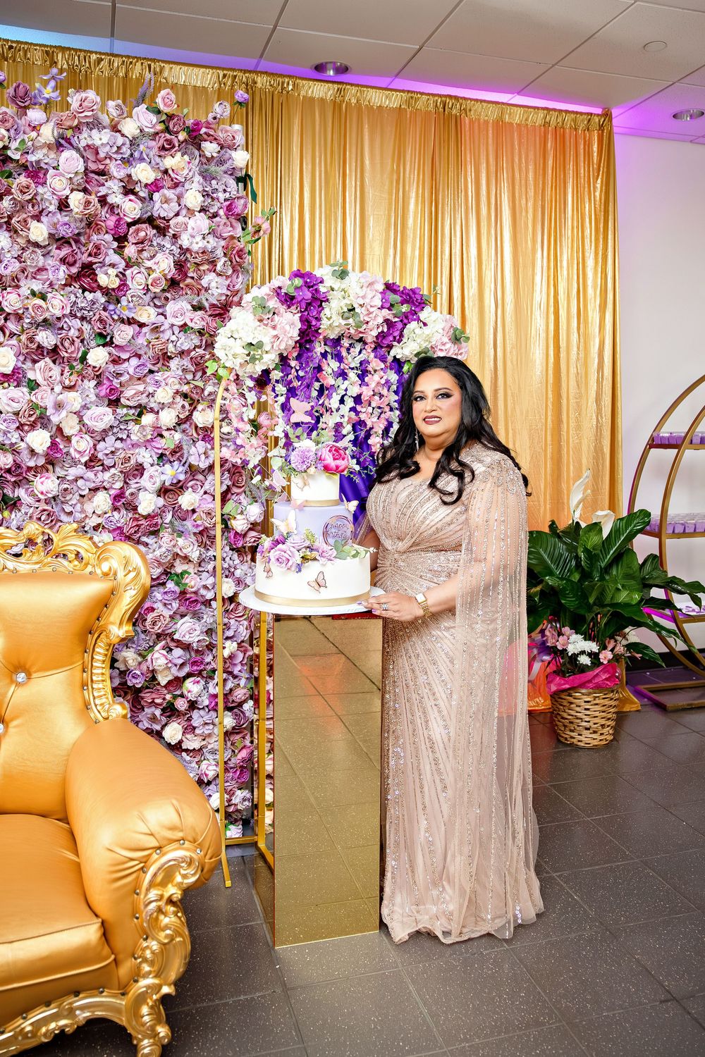 Woman in elegant gown poses with cake at floral backdrop celebration event with gold decor.
