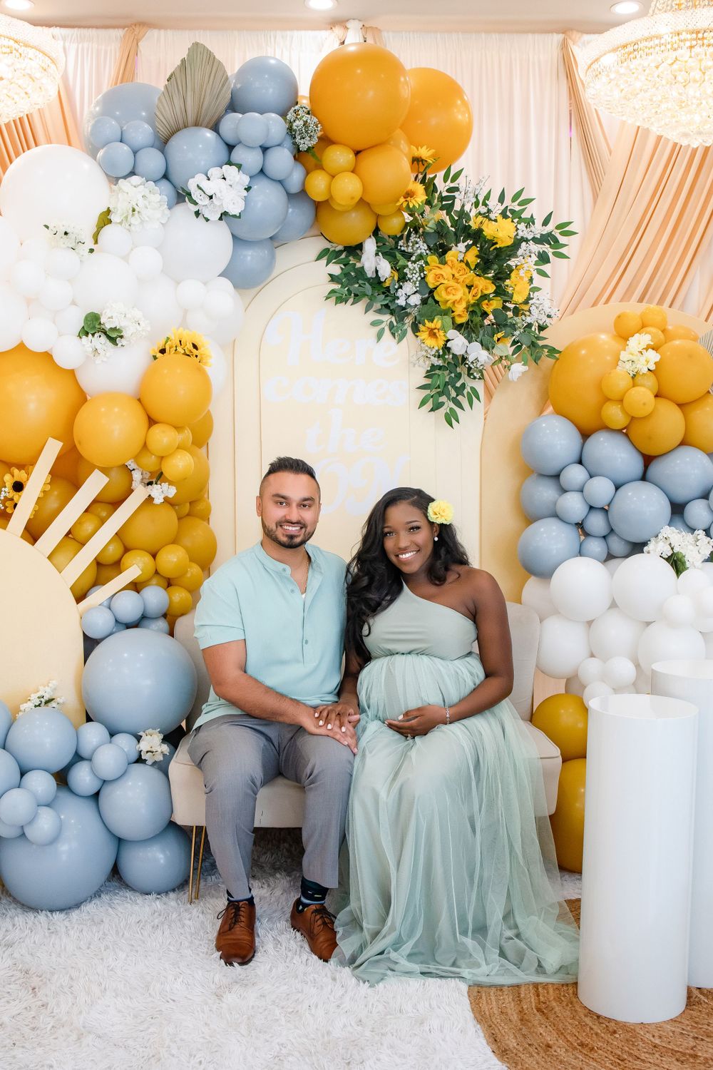 A pair stands on white carpet against a decorative balloon arch with yellow and blue accents.