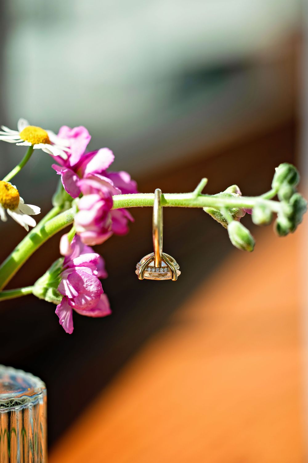 Gold engagement ring with gemstone hanging on a pink flower stem in a vase, close-up macro photography.