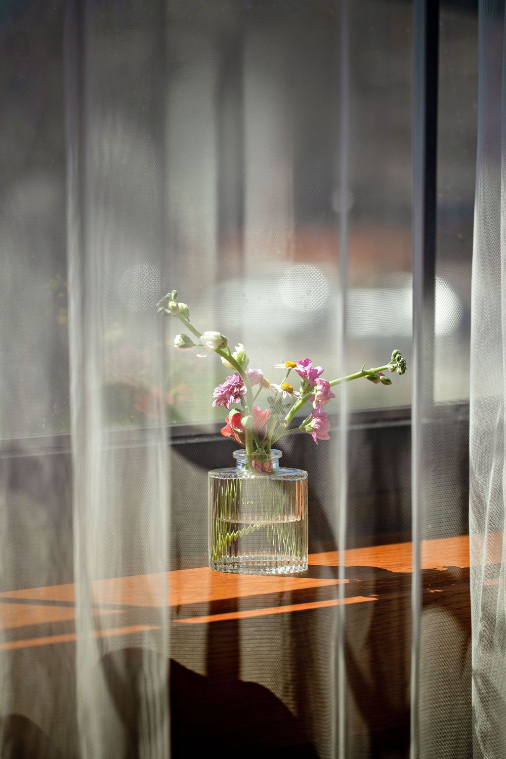 Small glass vase with pink and white wildflowers on a wooden windowsill, seen through sheer white curtains in soft sunlight.