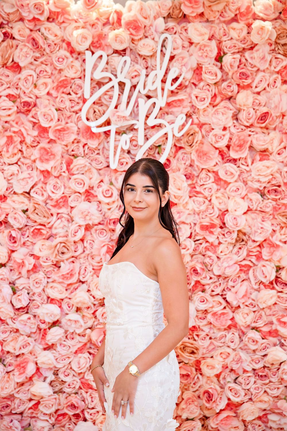 Bride-to-be in white strapless dress posing in front of pink rose flower wall with neon Bride to Be sign.