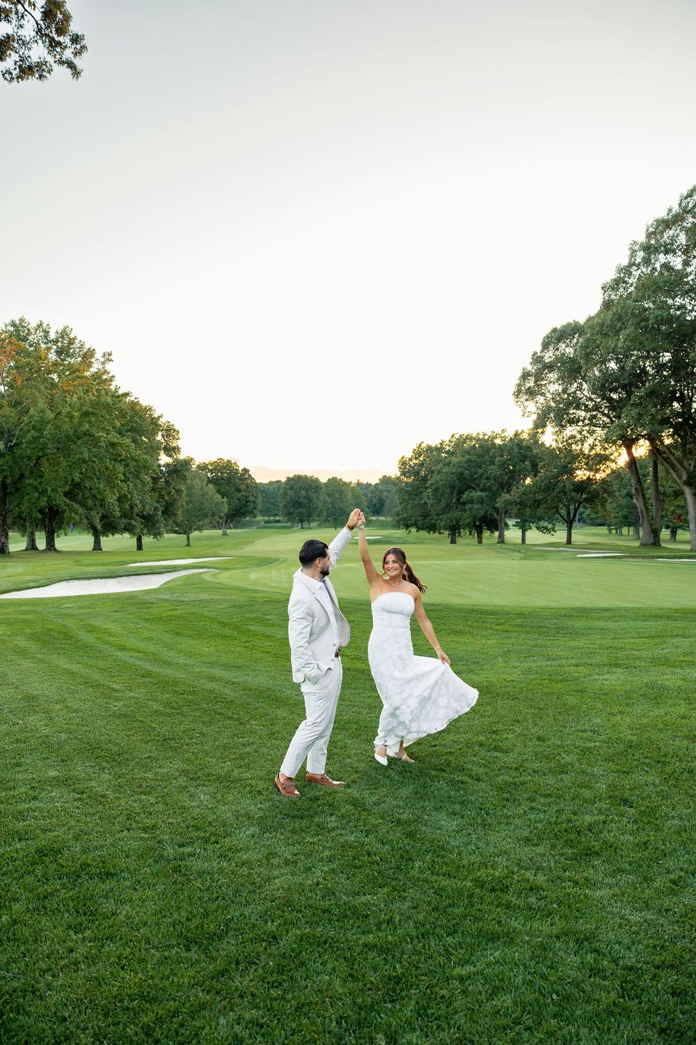 Bride and groom dancing joyfully on a lush green golf course at sunset, both dressed in white wedding attire.