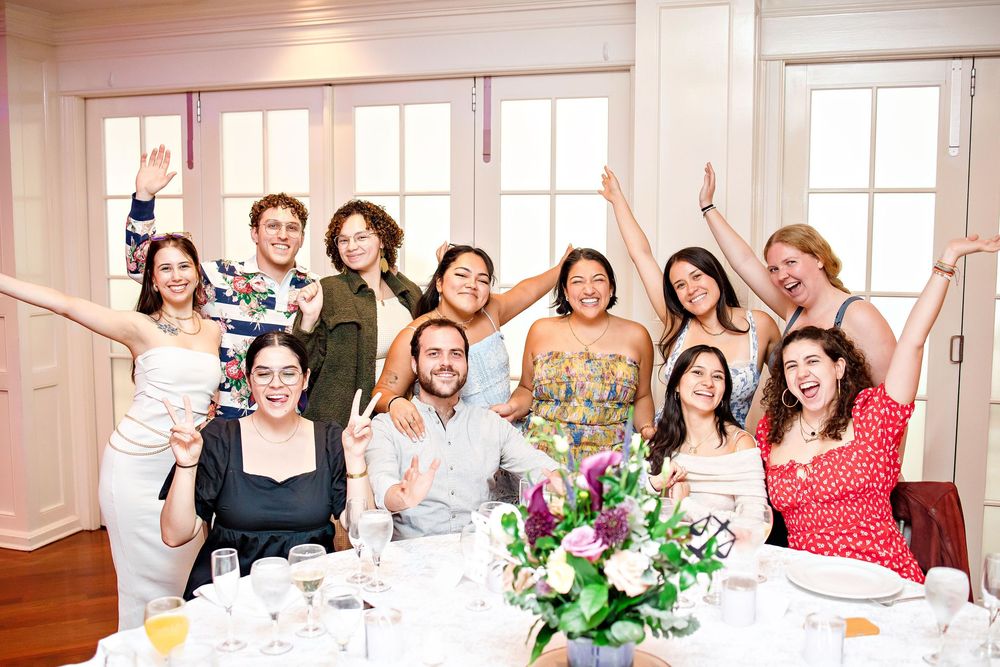 A group celebrates at a bright, sunny venue with a floral centerpiece on the dining table during a festive gathering.
