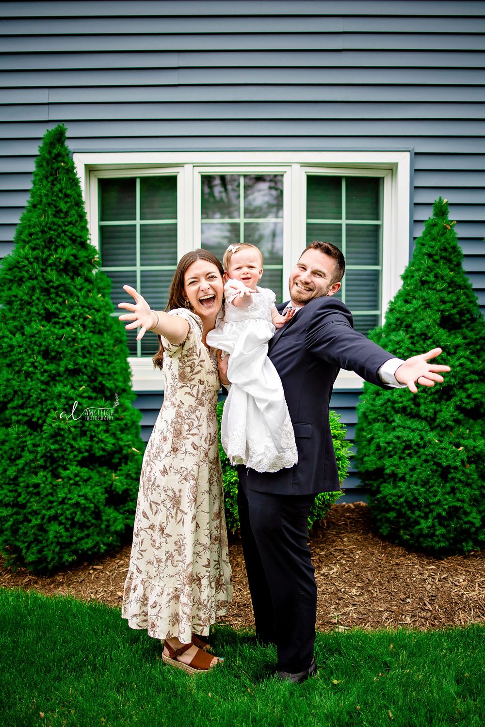 A happy family poses for a joyous outdoor photo in front of a house with manicured evergreen shrubs.