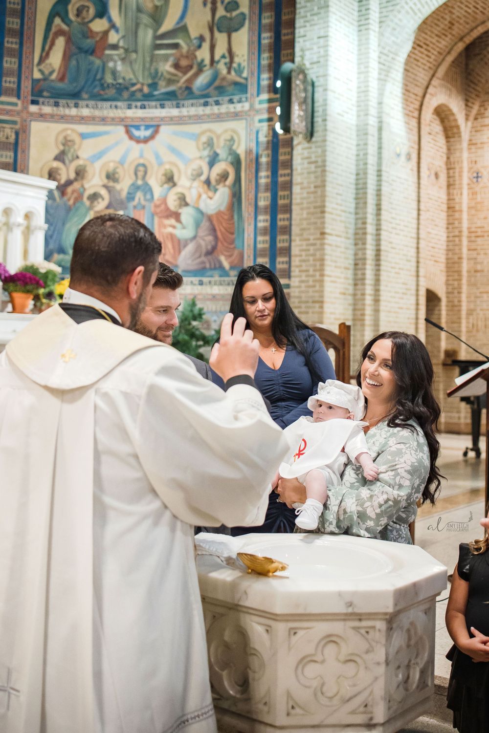 Baptism ceremony taking place at ornate stone font with religious murals in background.