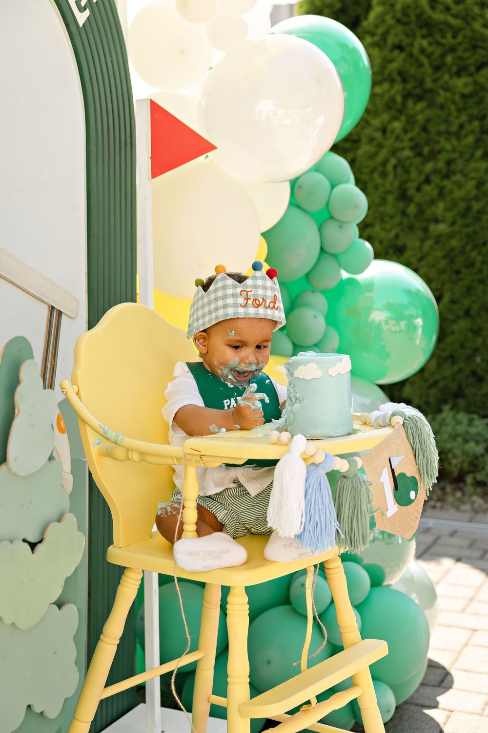 Toddler in birthday hat enjoys cake smash in yellow high chair, surrounded by green and white balloon decorations outdoors.