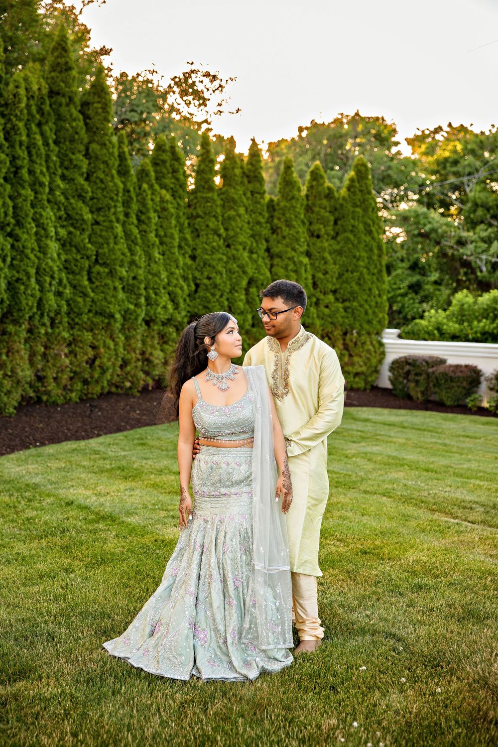 A couple in traditional South Asian attire poses on a lush green lawn surrounded by tall evergreen trees.