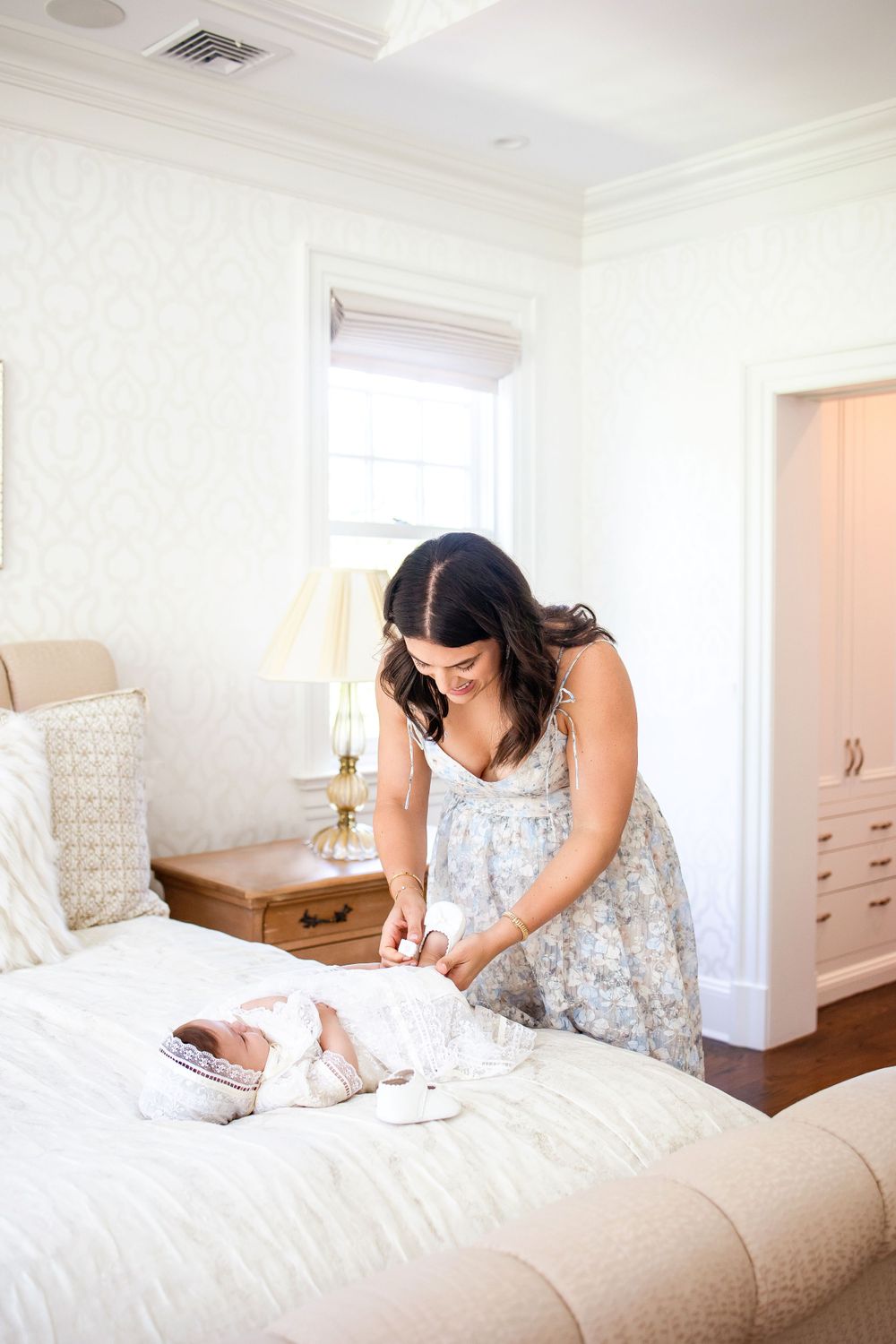 A woman in a floral dress arranges items on a white bed in a bright bedroom.