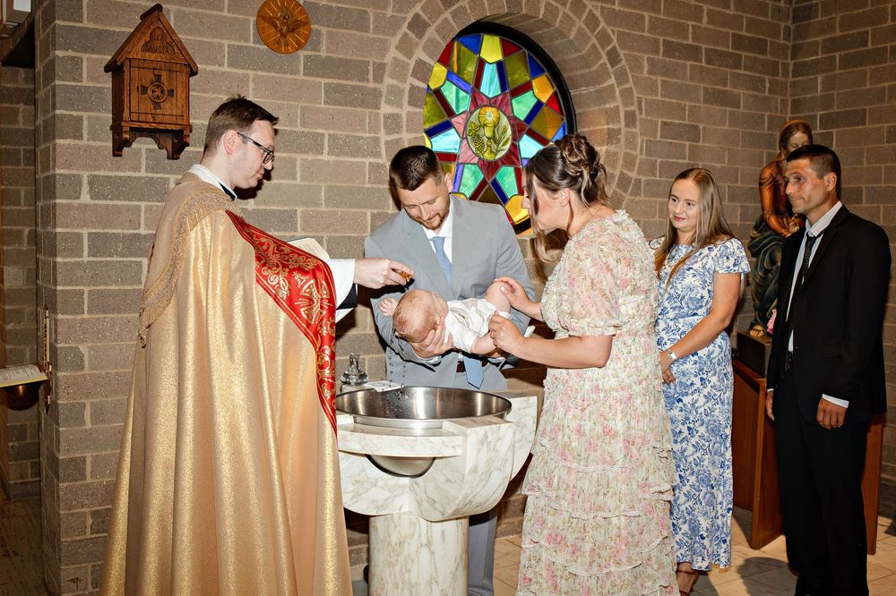 Priest performing a baptism ceremony at a stone font with parents and godparents in a church with stained glass.