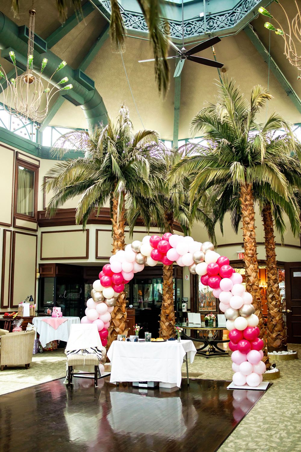 An elegant venue hall decorated with tall palm trees and pink and white balloon arches creates a romantic atmosphere.