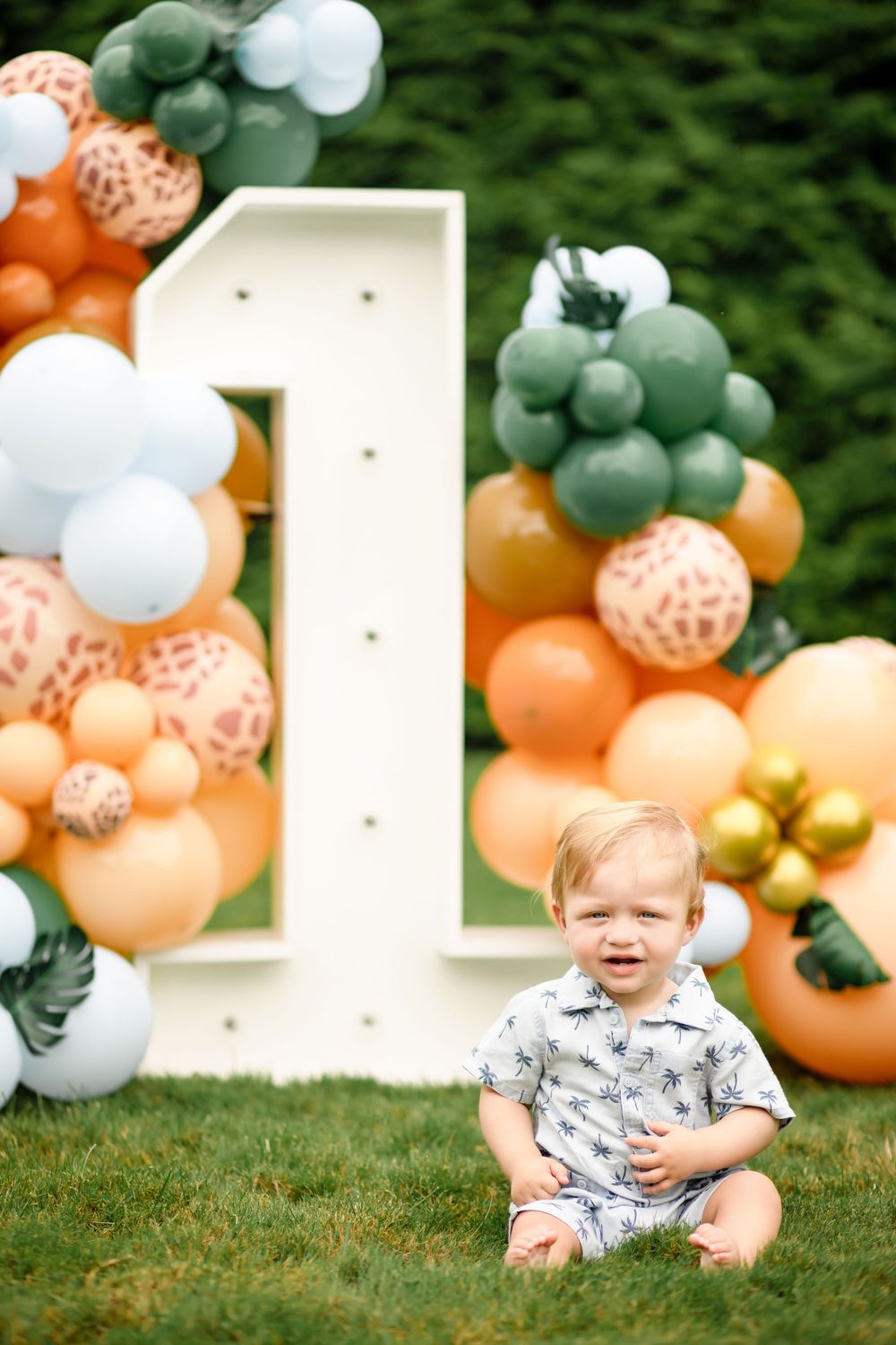First birthday celebration setup with large number one and balloon arch in orange, white and sage green colors.