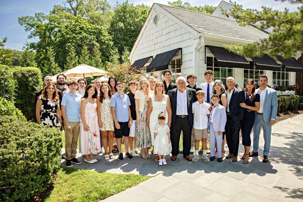 Large family group portrait outdoors near a charming white cottage, dressed in elegant attire for a special celebration.