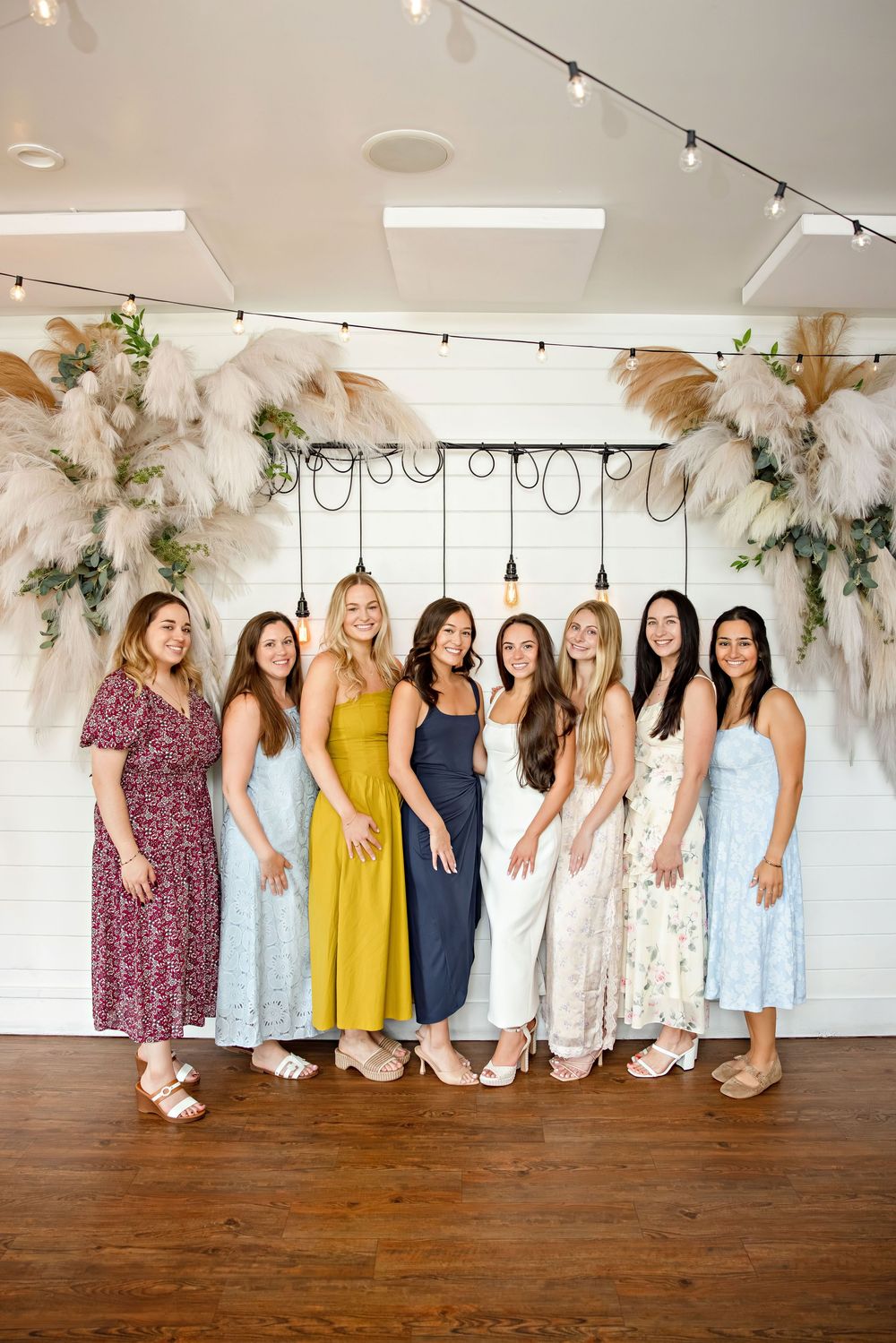 Eight women in colorful dresses posing together at an elegant indoor event venue with pampas grass decor.
