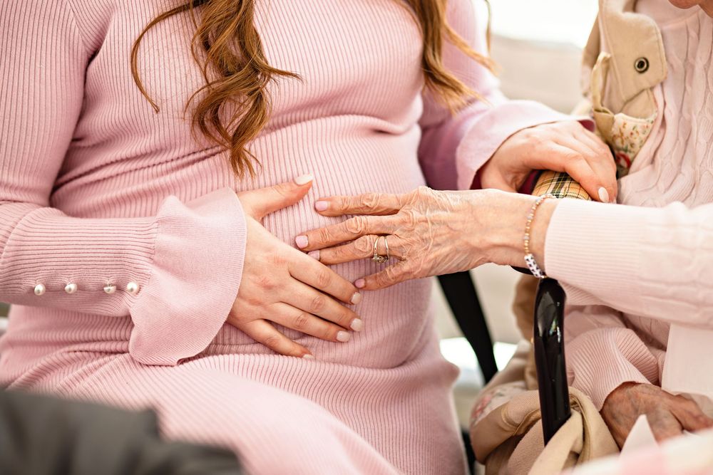 Elderly woman and pregnant woman in pink holding hands warmly, symbolizing generational bond and family connection.