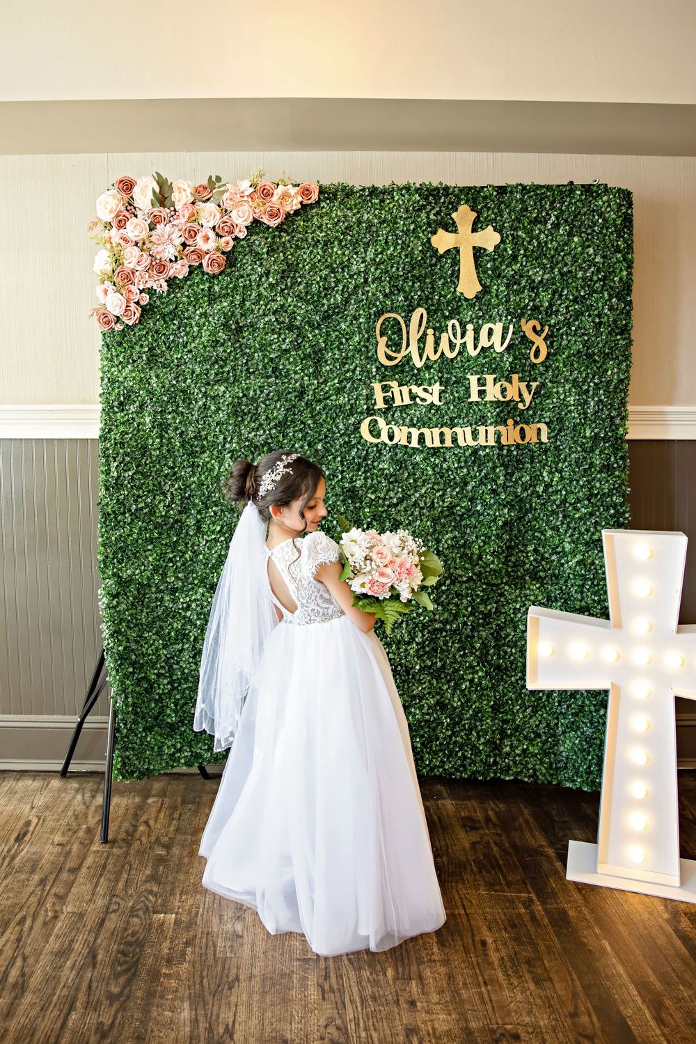 Girl in white dress and veil holds bouquet at Olivia's First Holy Communion celebration with floral green backdrop.