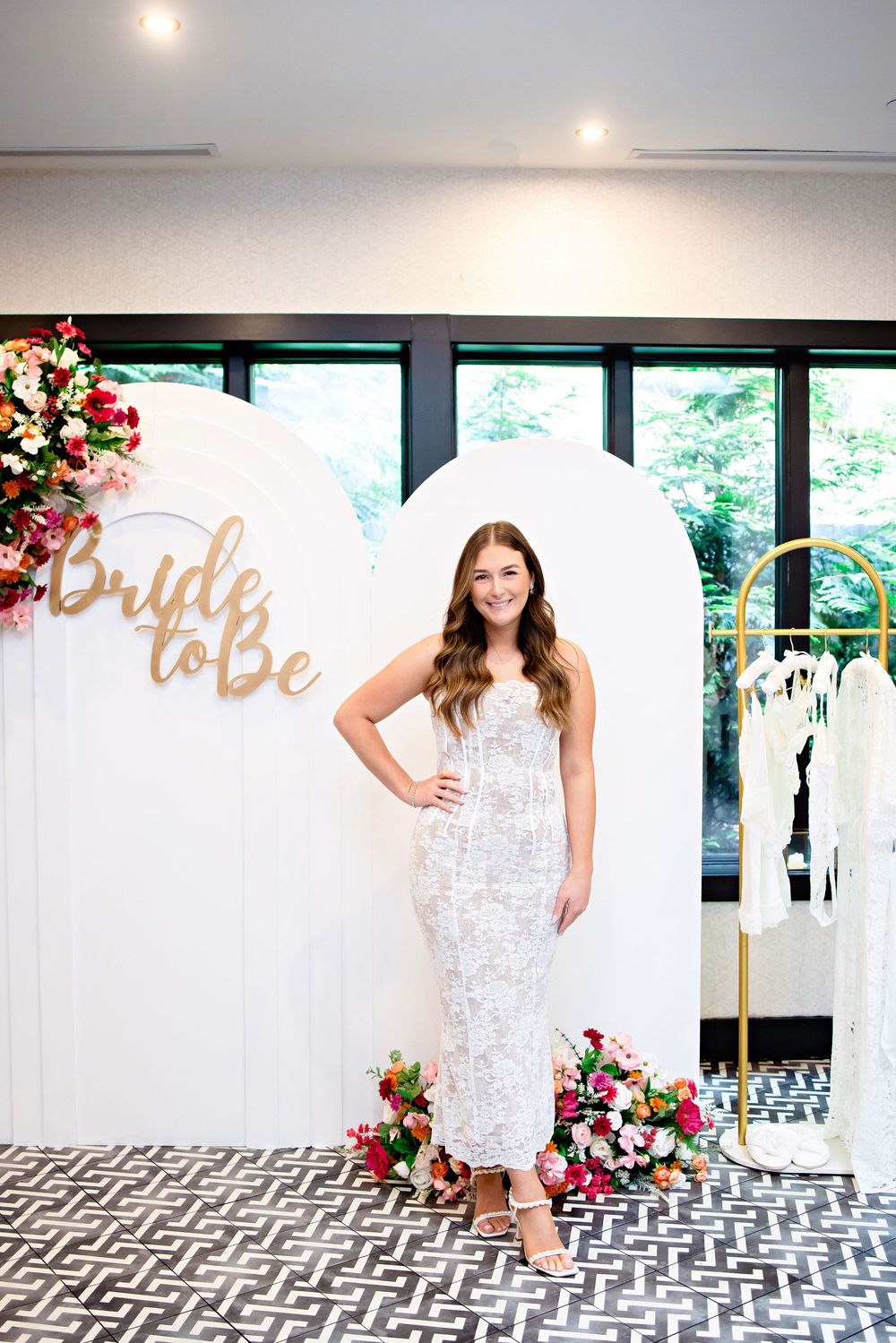 Elegant bridal fitting room with white decor, gold 'Bride to be' sign, geometric floor tiles and floral arrangements.