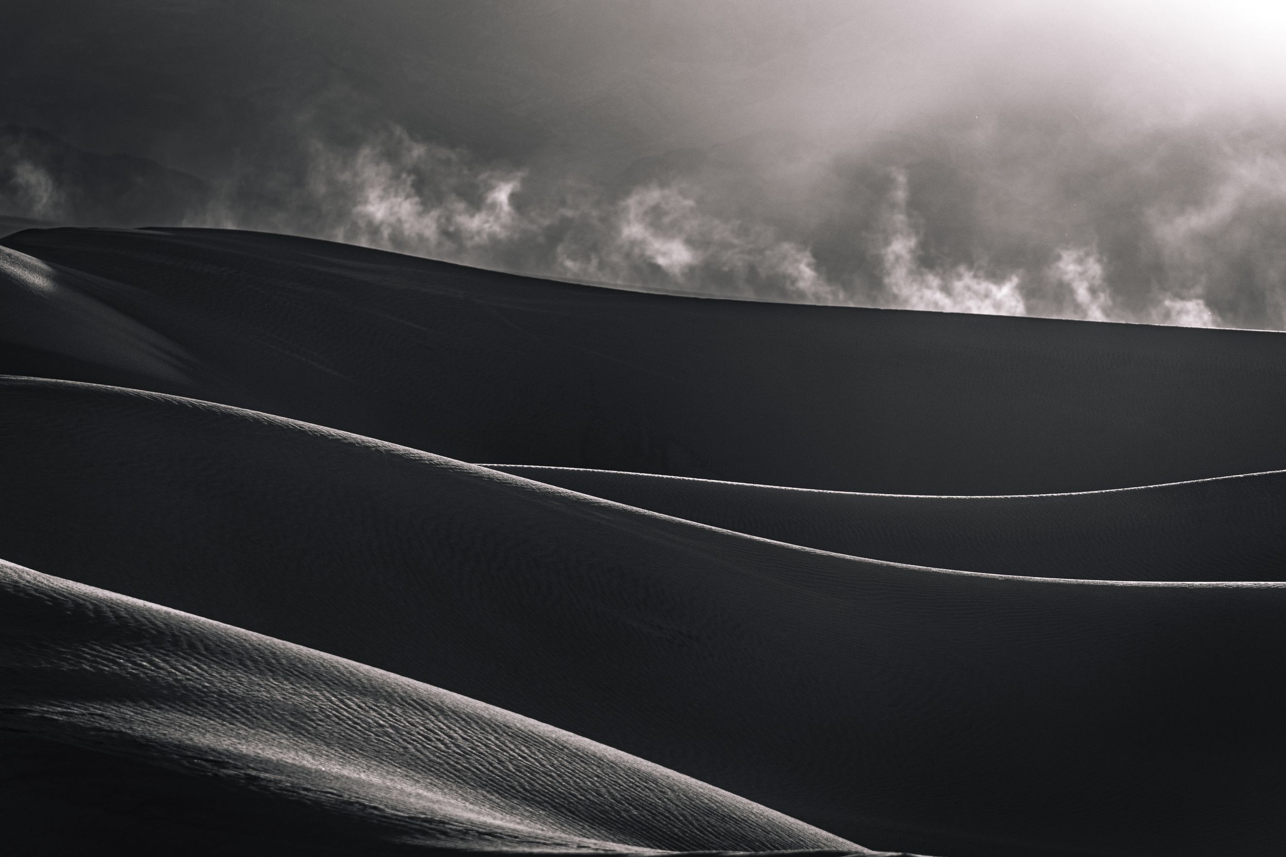 Black-and-white desert sand dunes, with a dramatic dust stream backlit, creating a striking contrast of light and shadow.