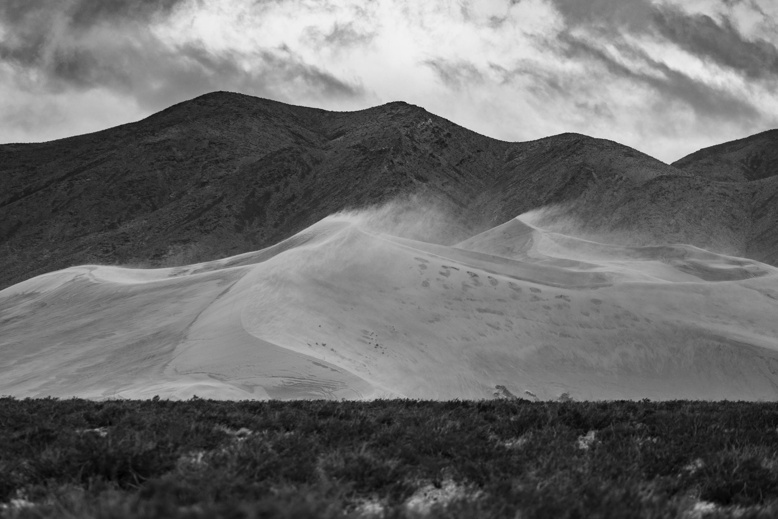 Black and white panoramic view of massive sand dunes and rugged mountains under cloudy skies.