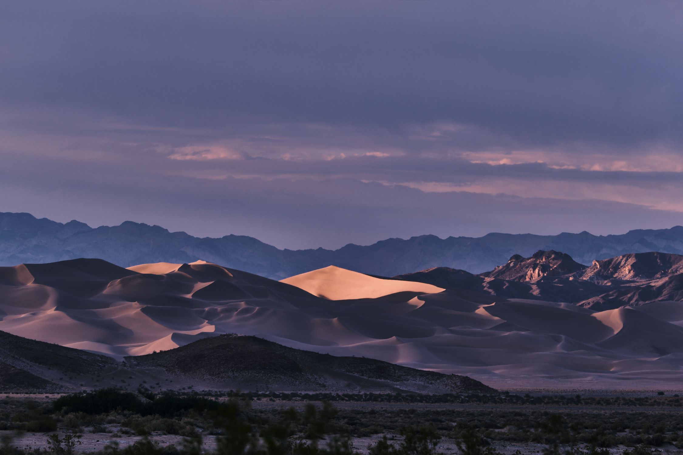 Purple dusk sky over illuminated sand dunes and silhouetted mountains in Death Valley desert.