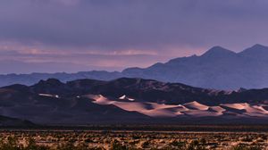 Sand dunes glow at dusk with layered mountain silhouettes under purple stormy sky.