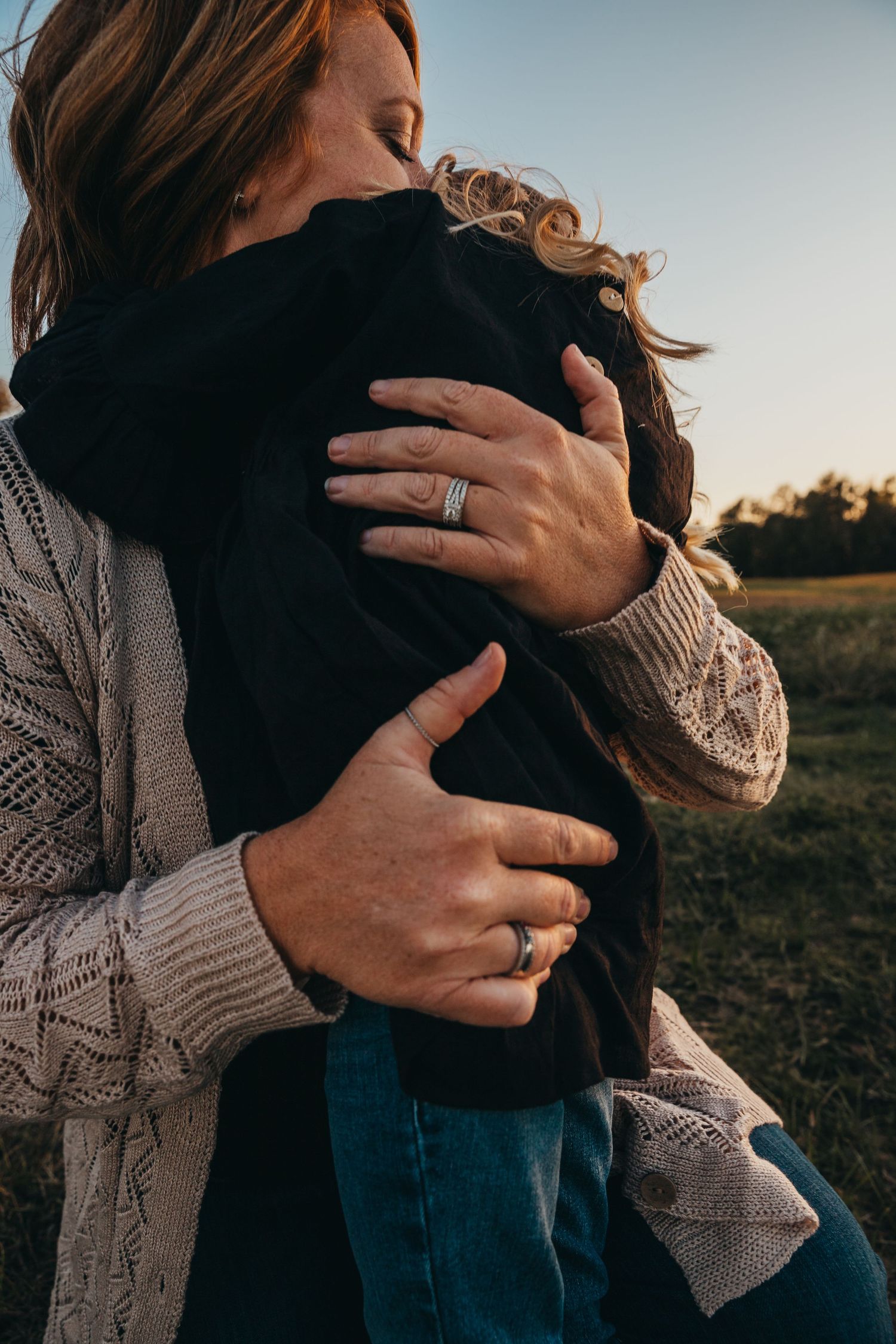 Person in knit sweater embracing another in outdoor setting during golden hour.