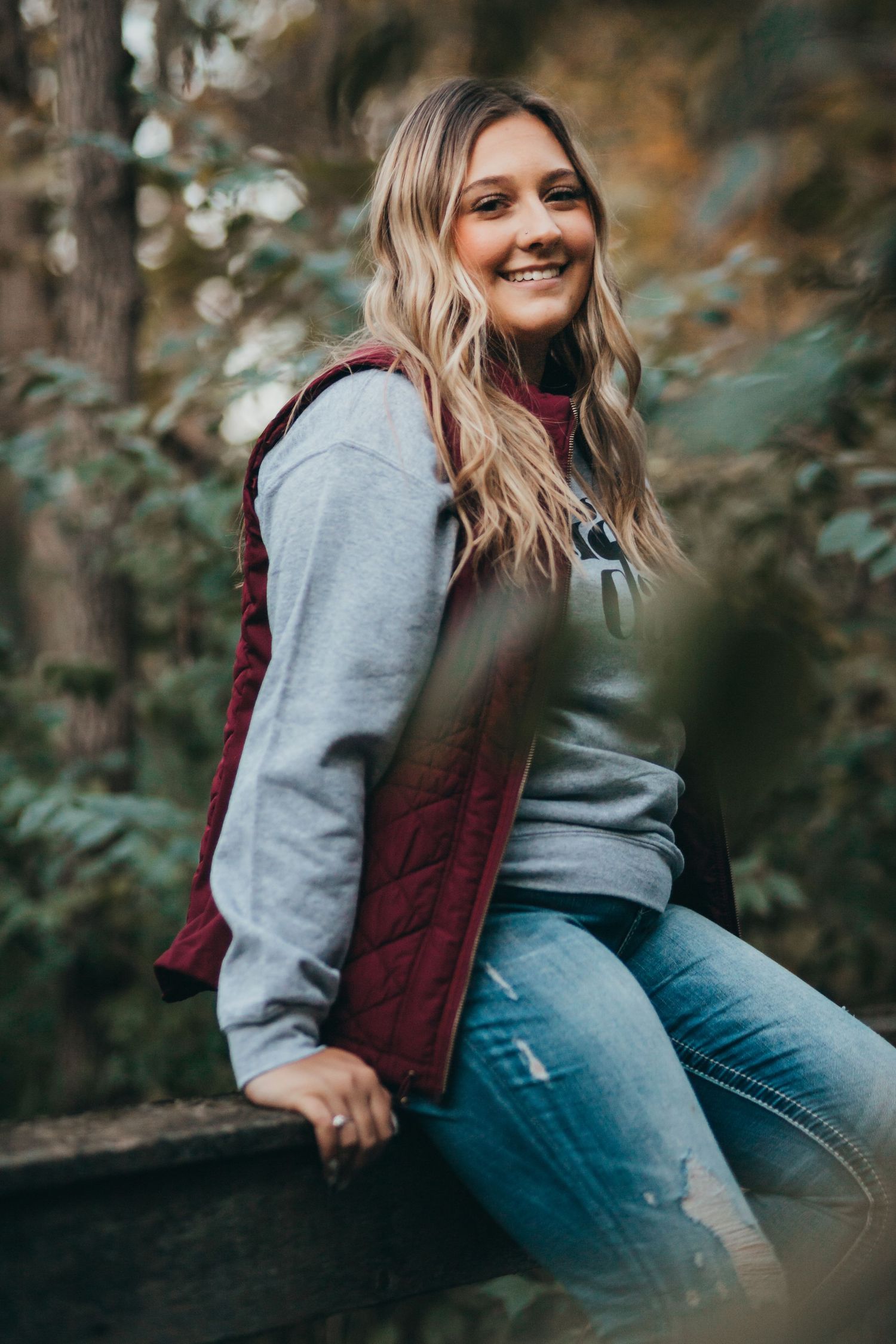 A person in casual autumn attire sits outdoors wearing a burgundy vest and grey sweater against a natural background.