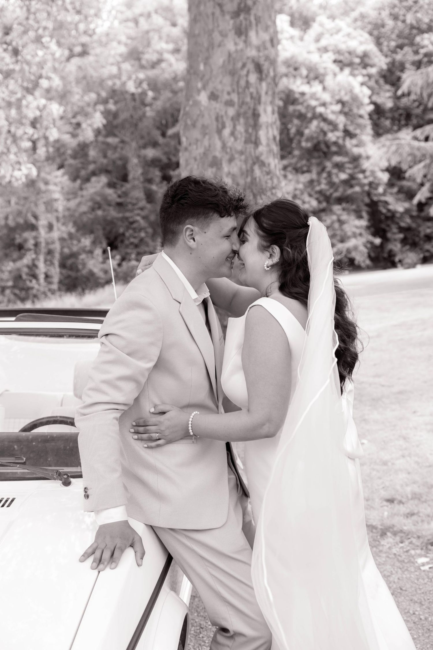 A romantic black and white moment captured beside a vintage car on a wedding day.