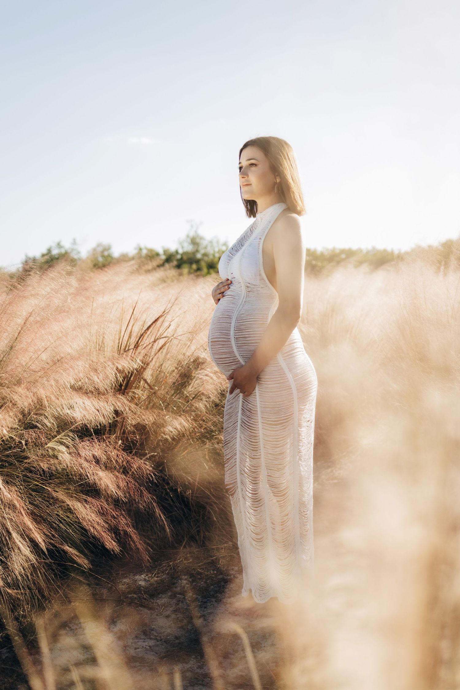 Ethereal maternity portrait of woman in white dress standing among sunlit tall grass.