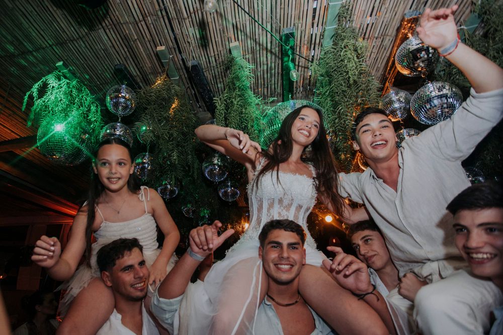 A group of party-goers dancing and celebrating at a nightclub with green lighting and disco balls in the background.