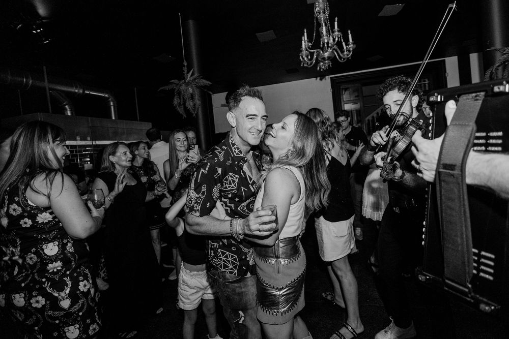 Couple dancing closely at a lively bar event with a violinist performing in the background.