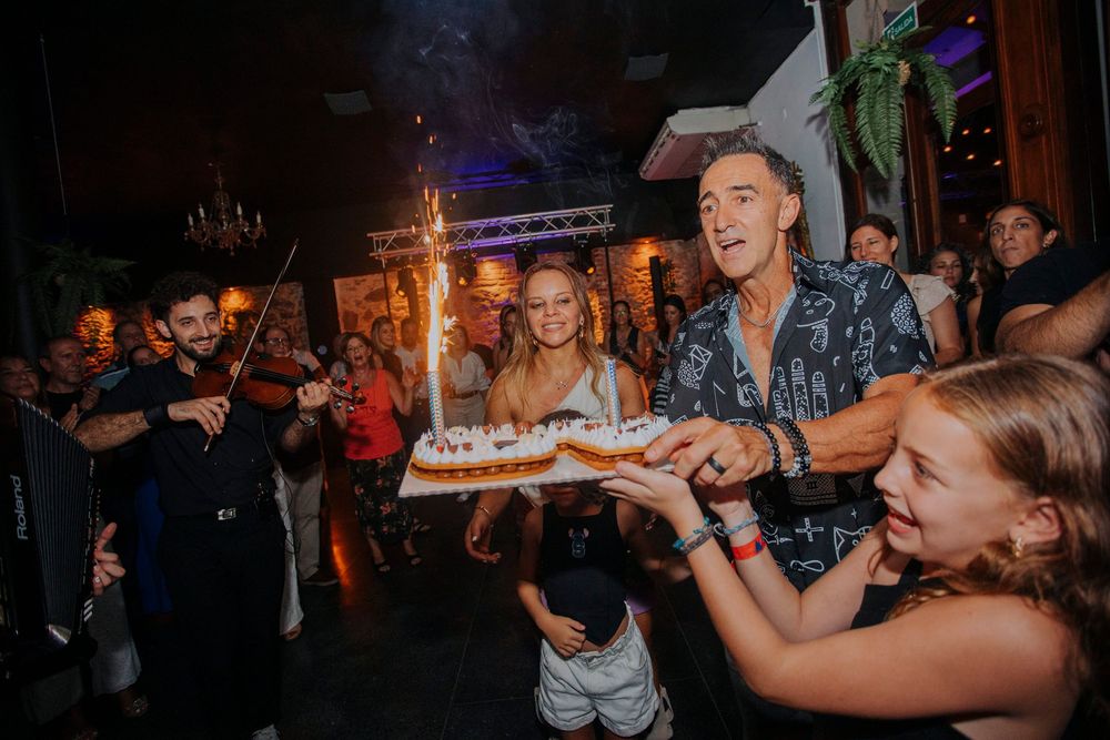 A man and woman celebrate a birthday with a sparkler cake surrounded by guests at a lively indoor party.