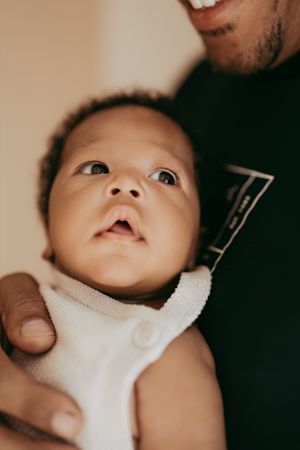 A baby wearing a white bib looks up with wide-eyed wonder while being held by someone in a green shirt.