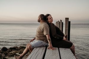 Two people sit back-to-back on a wooden dock overlooking the ocean at sunset.