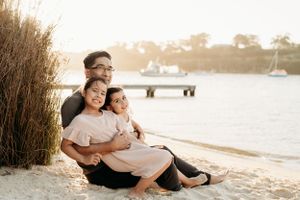 Family relaxing together on sandy beach with boats visible in background.