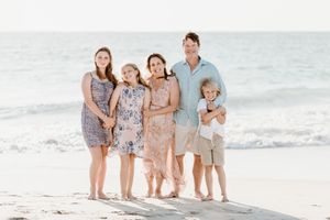 Family wearing coordinated pastel outfits poses together on a sunny beach with ocean waves in the background.
