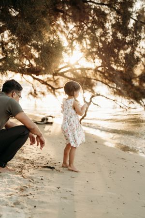 Child and adult exploring sandy beach under shaded trees during golden hour.
