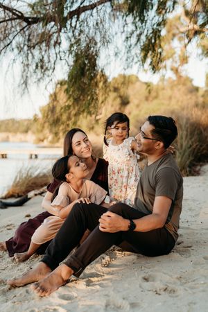 A family sits together on a sandy beach at sunset with weeping willow branches framing the scene.