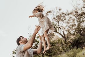 Parent playfully lifting child in the air against a bright sky backdrop.