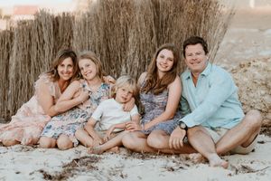 A family wearing coordinated neutral-colored outfits poses together on a sandy beach at sunset.