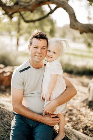 A parent and young child in a white dress pose together outdoors under a tree branch in warm sunlight.