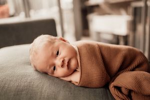 Baby wearing brown sweater lies peacefully on grey cushion while sleeping in sepia toned photograph.