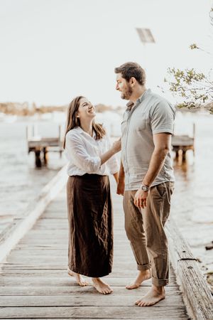 A couple shares a tender barefoot moment together while standing on a wooden pier by the water.