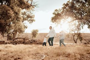 A couple walks their dog through a sun-drenched field with gnarled trees at golden hour.