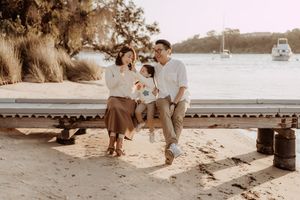 A couple sits together on a wooden bench by the beach during a romantic sunset photoshoot.